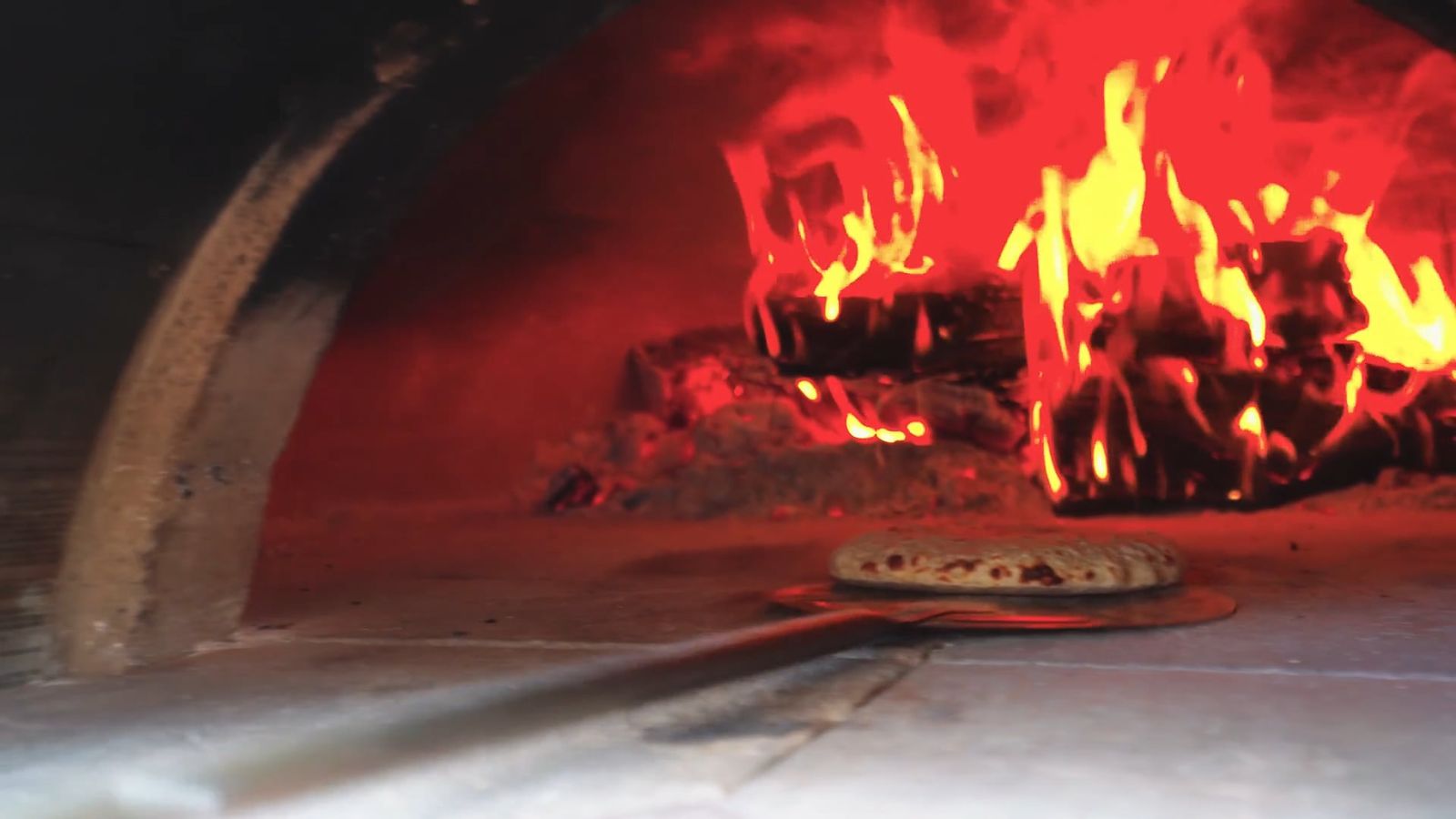Une pizza est enfournée dans un four à bois, les flammes au fond
