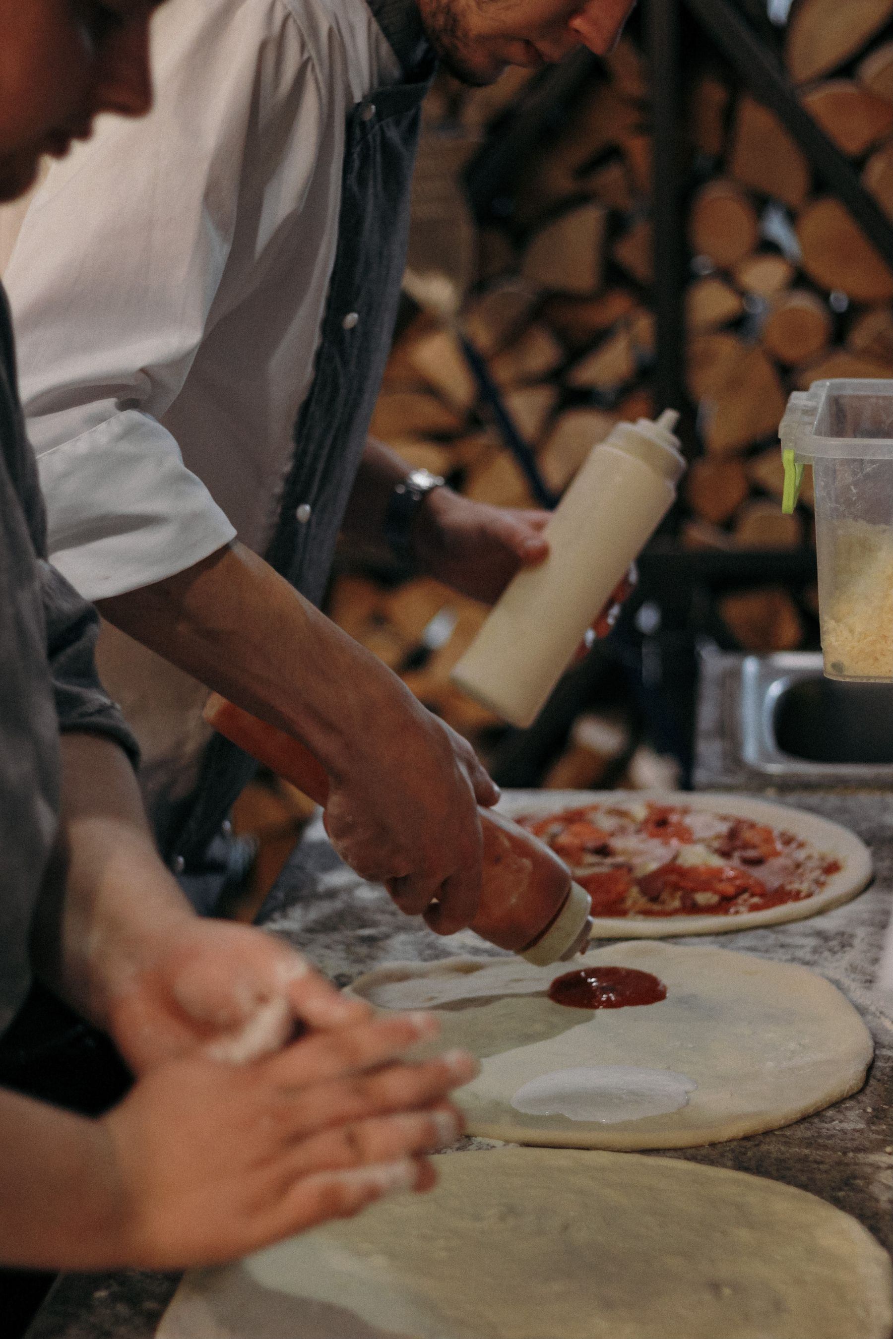 Un pizzaiolo garnit une pizza devant une pile de bûches